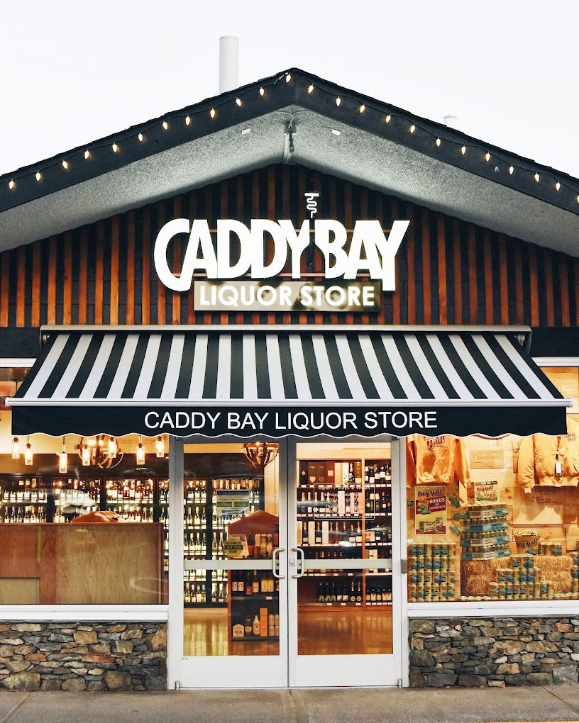 Cascadia Liquor Cabby Bay storefront with a sign that reads "Caddy Bay Liquor Store," featuring a striped awning and a well-lit display of bottles inside.